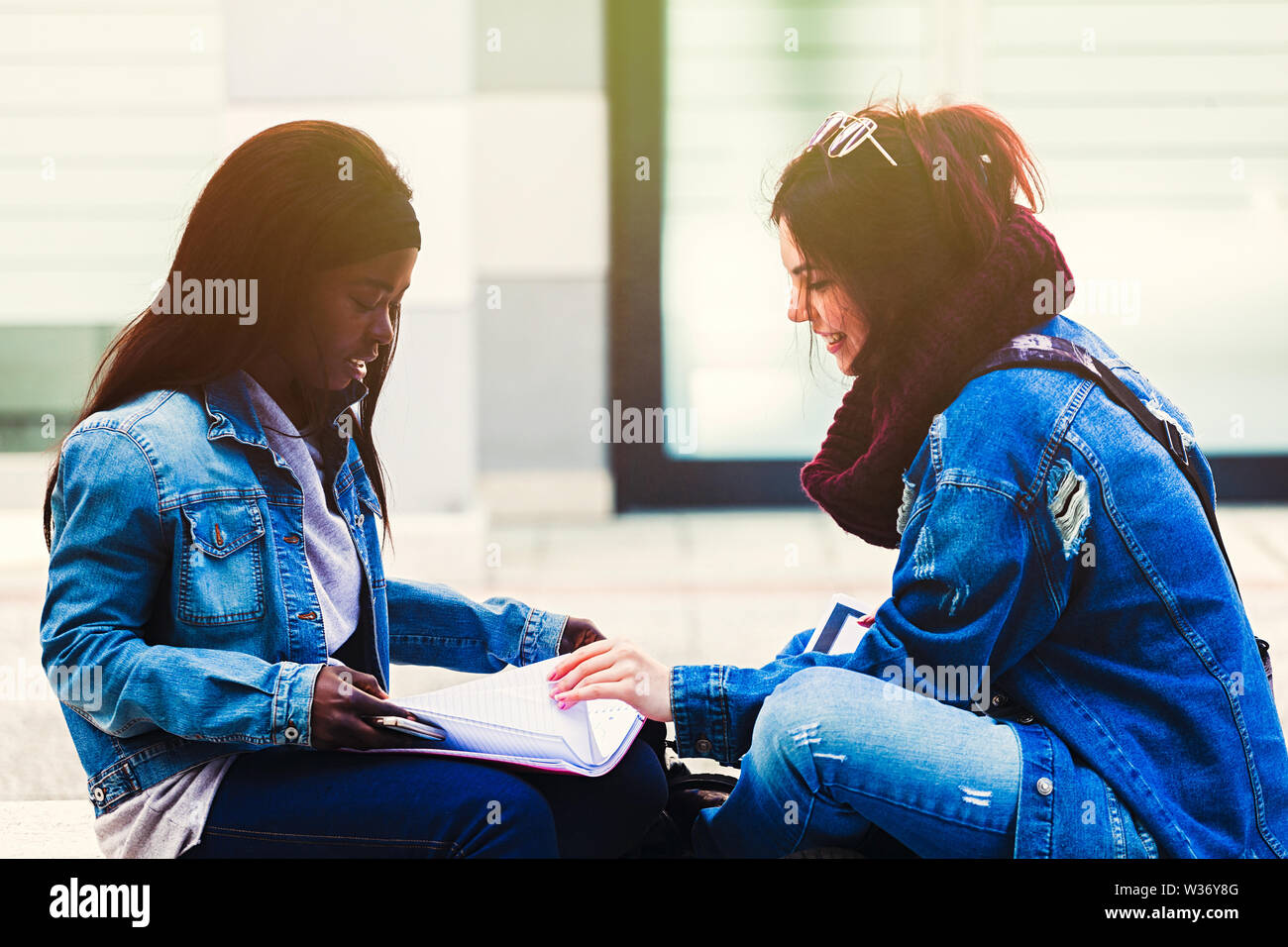 Due giovani donne, dell'Africano e Caucasico etnia, studio esterno seduta su una panchina. Foto Stock