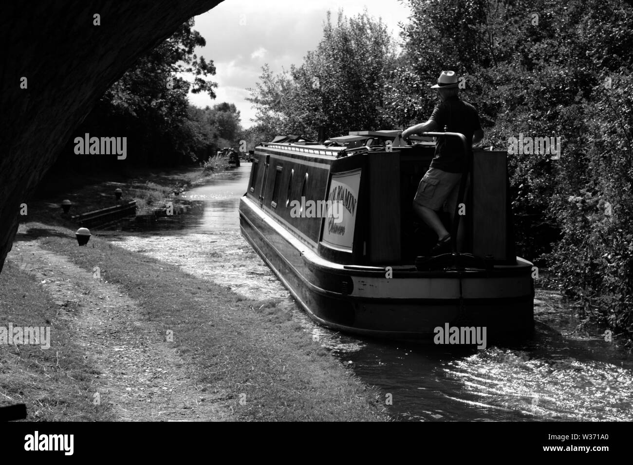 Barca stretta che viene guidato da un uomo che indossa un cappello da sole sul Canal Grand Union subito dopo aver lasciato una serratura abbassata, vicino Aylesbury, Buckinghamshire. Foto Stock
