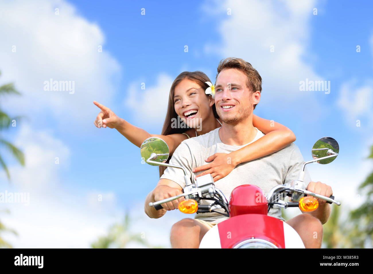 Felice coppia dispositivo di puntamento su scooter travel vacation. Giovane donna asiatica che mostra il suo fidanzato di una posizione durante la guida di un ciclomotore su un viaggio durante le vacanze estive con cielo blu e nuvole sfondo. Foto Stock