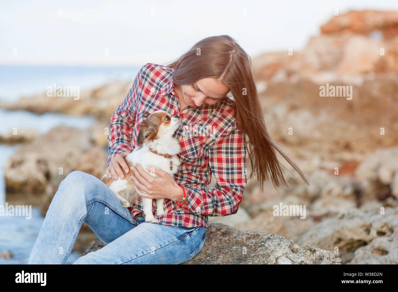 Piuttosto giovane donna adulta indossando plaid shirt e jeans seduti sulla costa del mare mentre tiene il suo piccolo cane chihuahua cercando nei suoi occhi Foto Stock