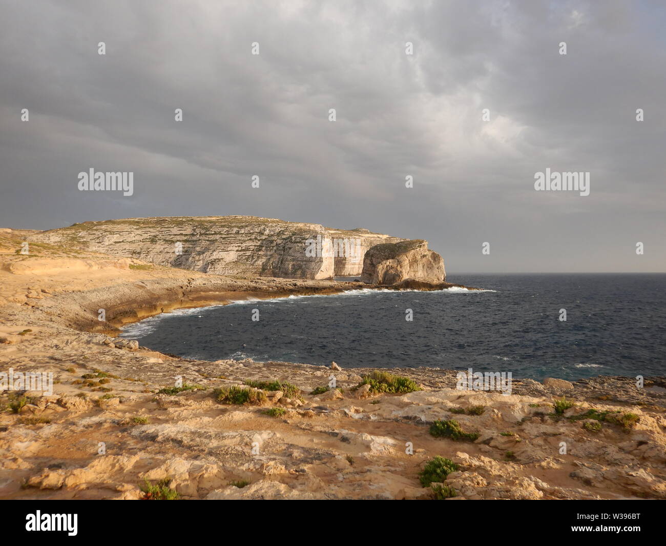 Bella round bay, piante verdi e Fungus Rock Cliffs al tramonto a Gozo. Arancione della luce al tramonto con il grigio nuvole temporalesche. Foto Stock