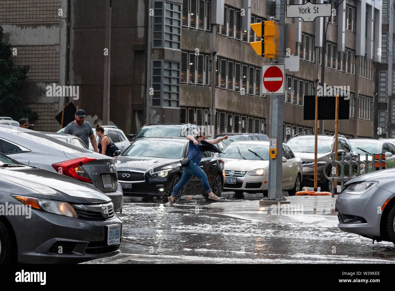 Toronto, Canada. Luglio 13, 2019. Pesante pioggia del pomeriggio ha lasciato molti downtown intersezioni inondati di Toronto. Foto di una femmina di salto pedonale su acqua mentre si incrociano a York e Porto Street. Credito: EXImages/Alamy Live News Foto Stock