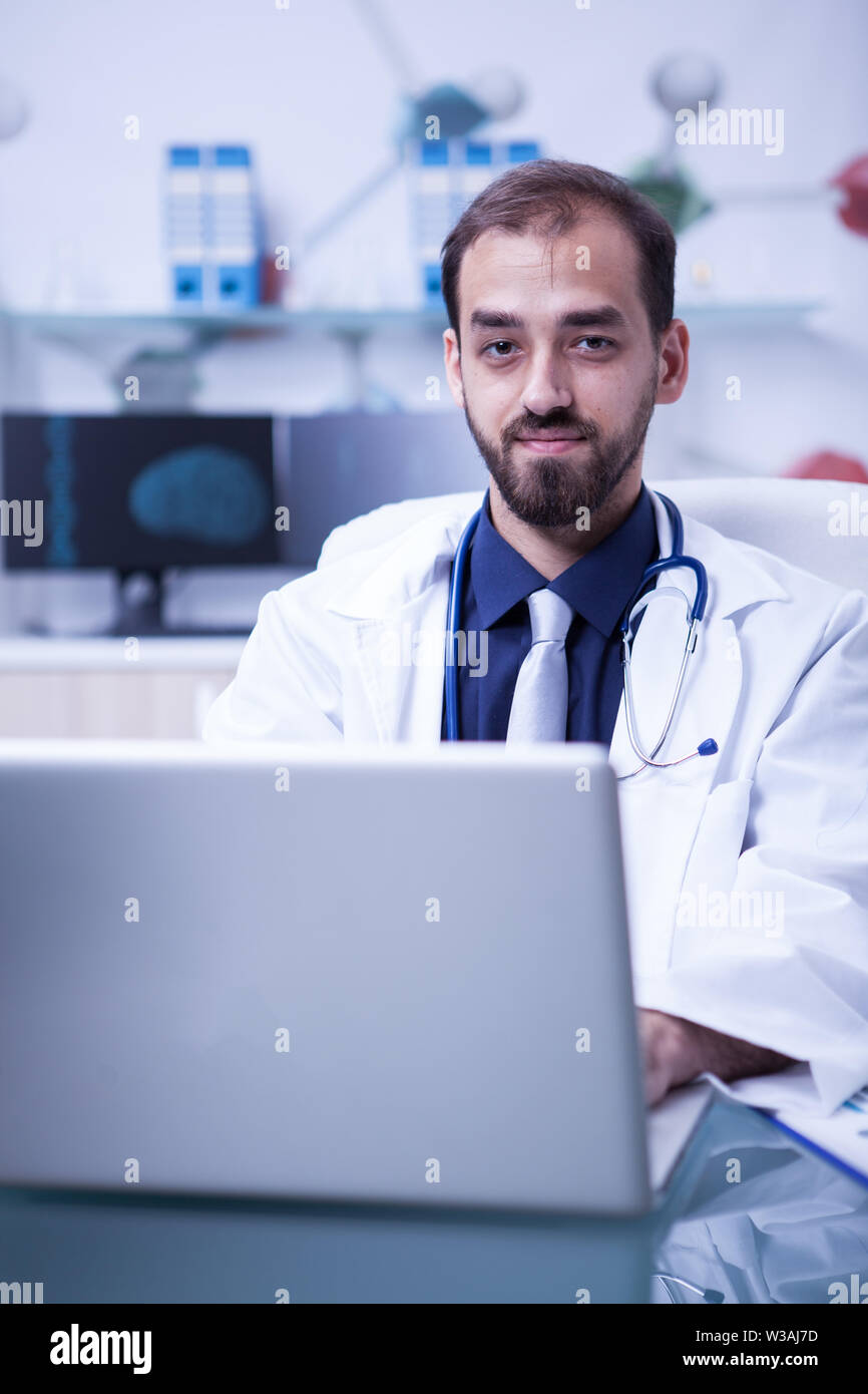 Ritratto di giovane medico che lavorano su laptop e sorridente tot la fotocamera. Medico con lo stetoscopio intorno al suo collo. Foto Stock