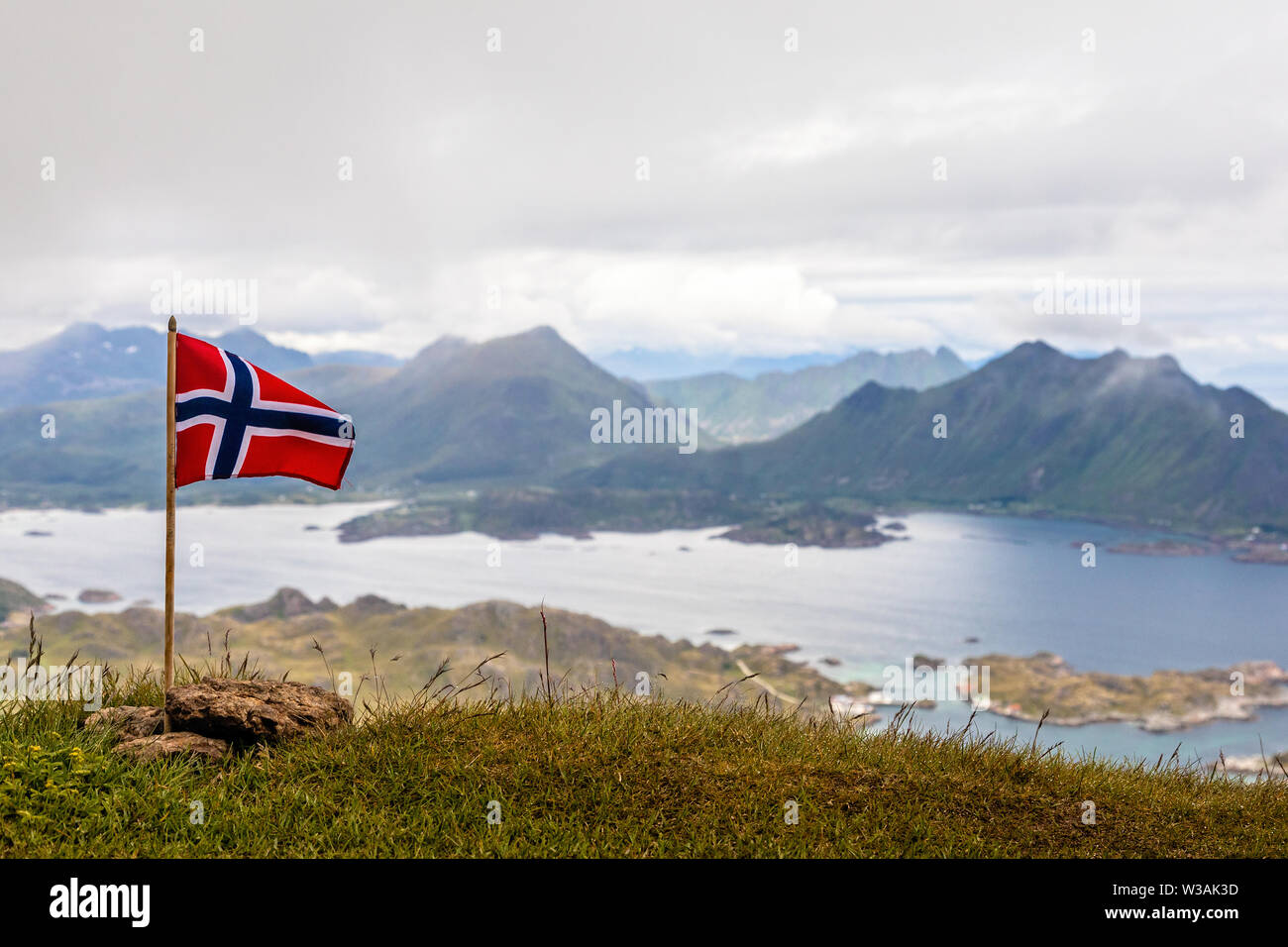 Norwegian bandiera nazionale rinuncia al vento sulla sommità del picco Nonstinden con fjord in background, Ballstad, Vestvagoy comune, Nordland cou Foto Stock