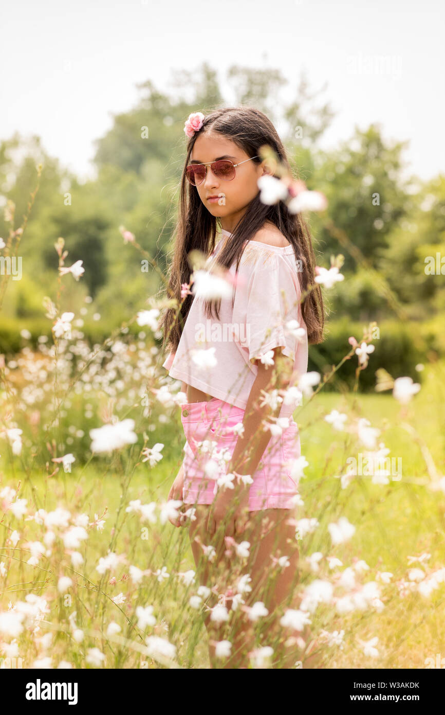 Ragazza giovane e carina in rosa moda estate indossando occhiali da sole, pantaloncini e top con fiore nei suoi capelli piedi tra fiori di campo in un campo rurale Foto Stock