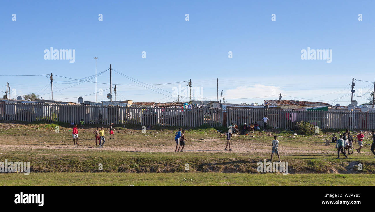 Gruppo di poveri neri africani i bambini giocando una partita di calcio o di calcio su un orlo di erba davanti a un recinto e baracche di stagno Cape Town, Sud Africa Foto Stock