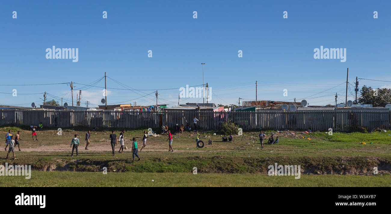 Gruppo di poveri neri africani i bambini giocando una partita di calcio o di calcio sul lato dell'autostrada con stagno shack case in background Cape Town Foto Stock