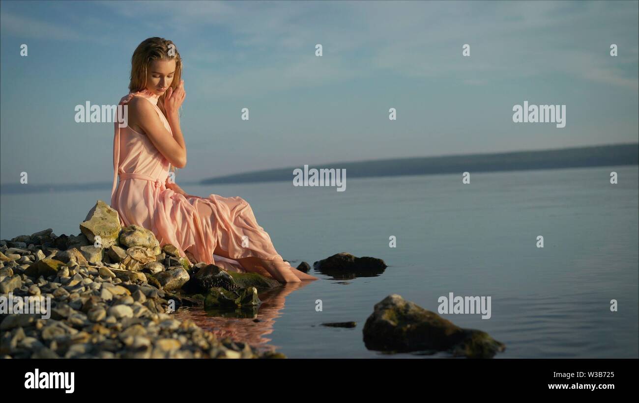 Lonely donna sogna e guardando alla bellissima alba sul molo con il mare e le montagne sullo sfondo. Vista posteriore, piano generale. La ragazza si siede in estate Foto Stock