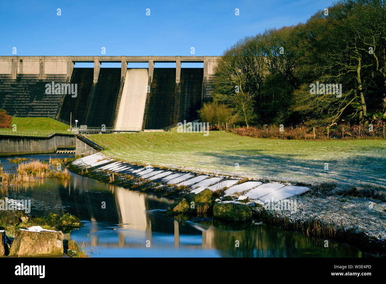 Alta e ripida, diga in calcestruzzo a serbatoio Thruscross riflessa nell'acqua che fluisce nel canale del gelido inverno giorno (blue sky) - North Yorkshire, Inghilterra, Regno Unito Foto Stock