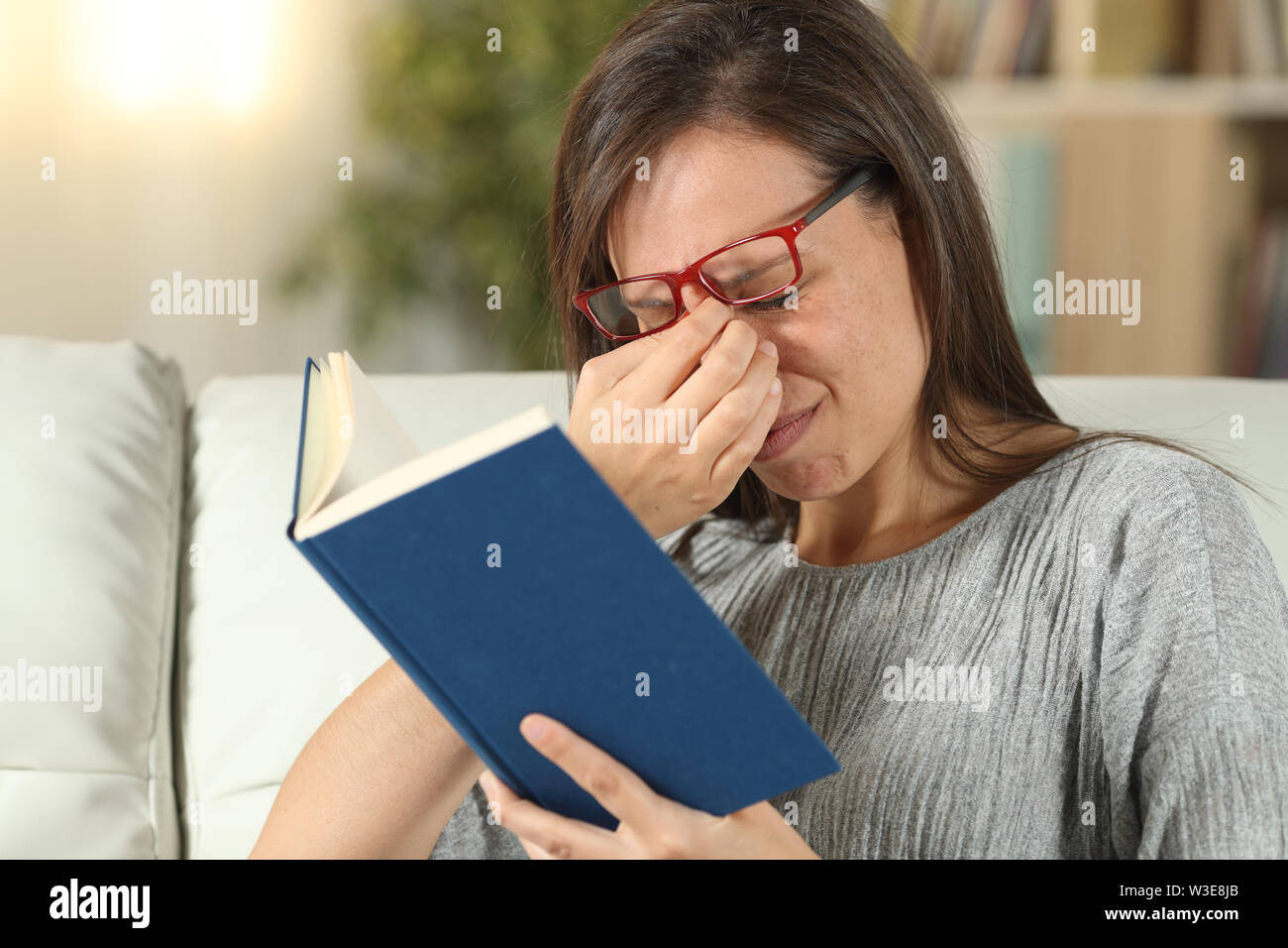 Donna con occhiali sofferenza affaticamento degli occhi la lettura di un libro seduti su un divano nel salotto di casa Foto Stock