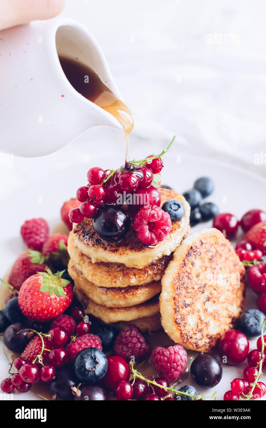 Donna versando lo sciroppo d'acero sul gustoso formaggio frittelle servite con i lotti dei frutti di bosco freschi su sfondo bianco. Pila di pancake con colata di miele Foto Stock