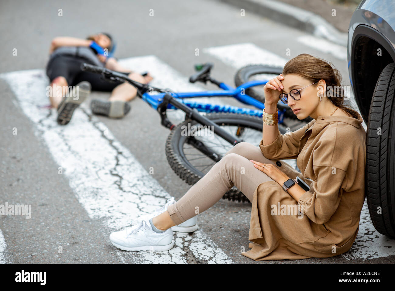 Triste donna seduta vicino alla vettura, preoccupato dell'uomo ferito giacente sul passaggio pedonale dopo l'incidente stradale Foto Stock