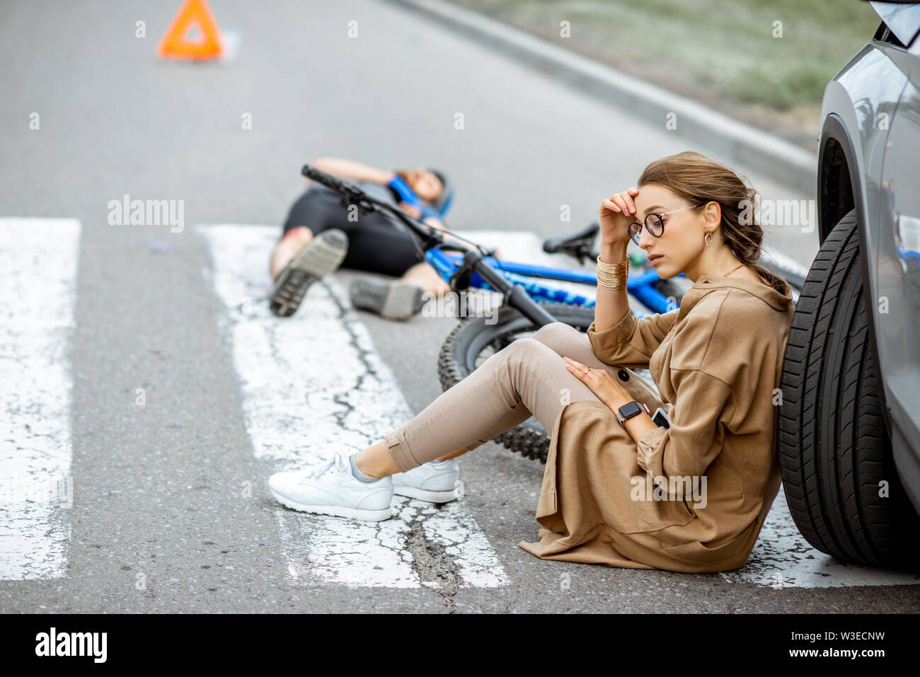 Triste donna seduta vicino alla vettura, preoccupato dell'uomo ferito giacente sul passaggio pedonale dopo l'incidente stradale Foto Stock