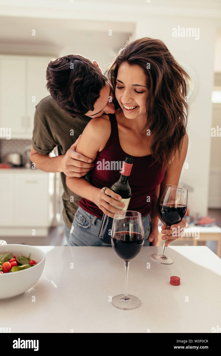 Giovane aventi data di notte. Uomo in piedi dietro una donna il riempimento di bicchieri di vino in cucina. Coppia romantica in cucina con il vino. Foto Stock