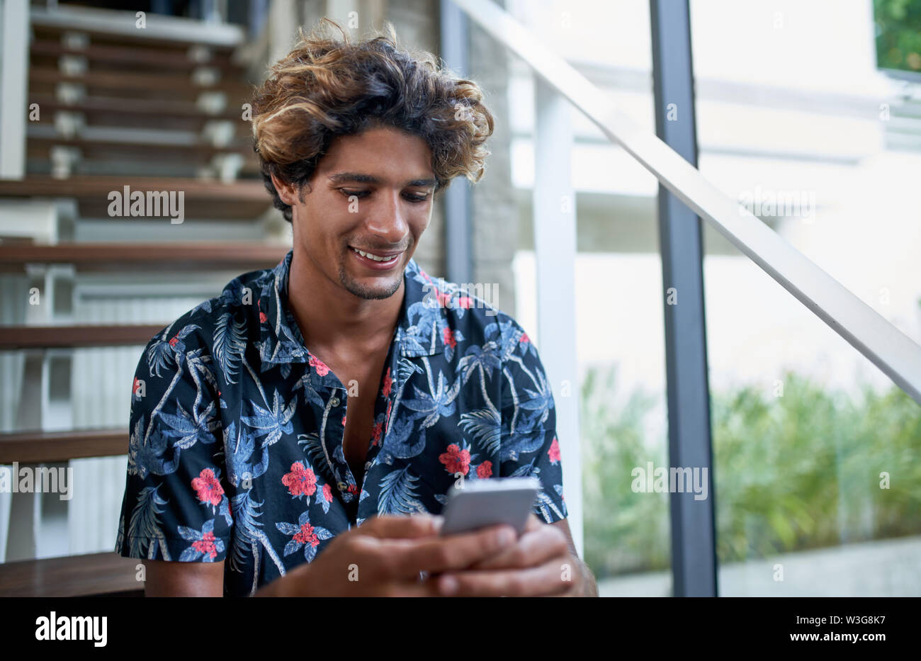 Stile di vita closeup colpo di bello brasiliano balinese millenial influencer in 20s, indossando variopinti camicia floreale seduti sulle scale del moderno e alla moda di un Foto Stock