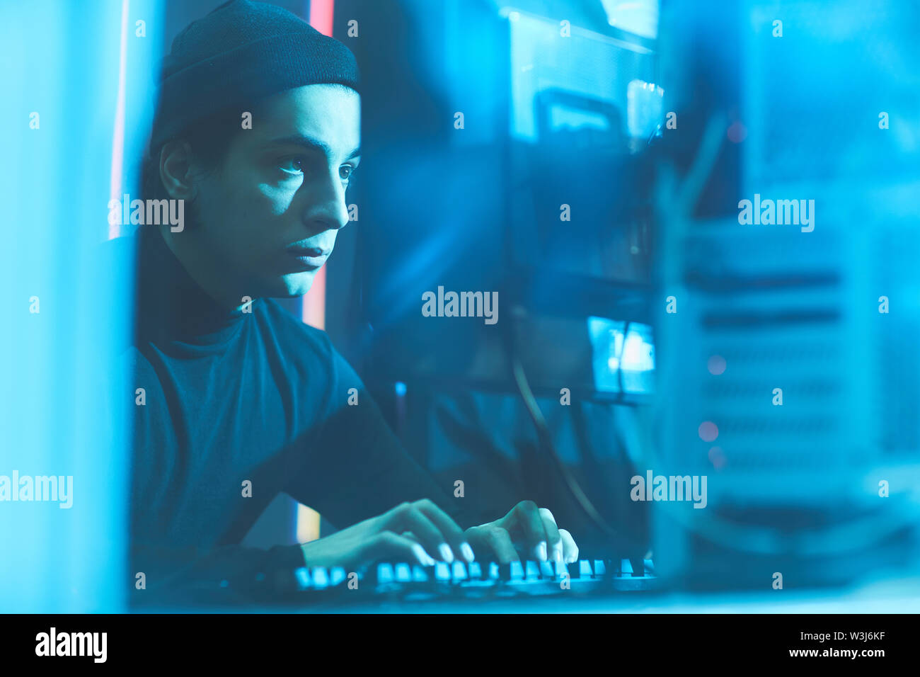 Grave concentrata giovani hacker ebraica in beanie hat seduta a tavola in camera oscura e digitando sulla tastiera mentre la rottura in dati server Foto Stock