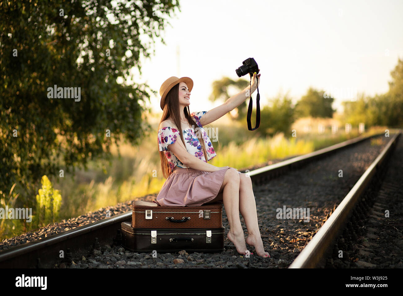 Un esile ragazza in un cappello di paglia che è seduto su una delle valigie sui binari della ferrovia, tenendo una fotocamera e scattare foto di se stessa. Il concetto di viaggio Foto Stock