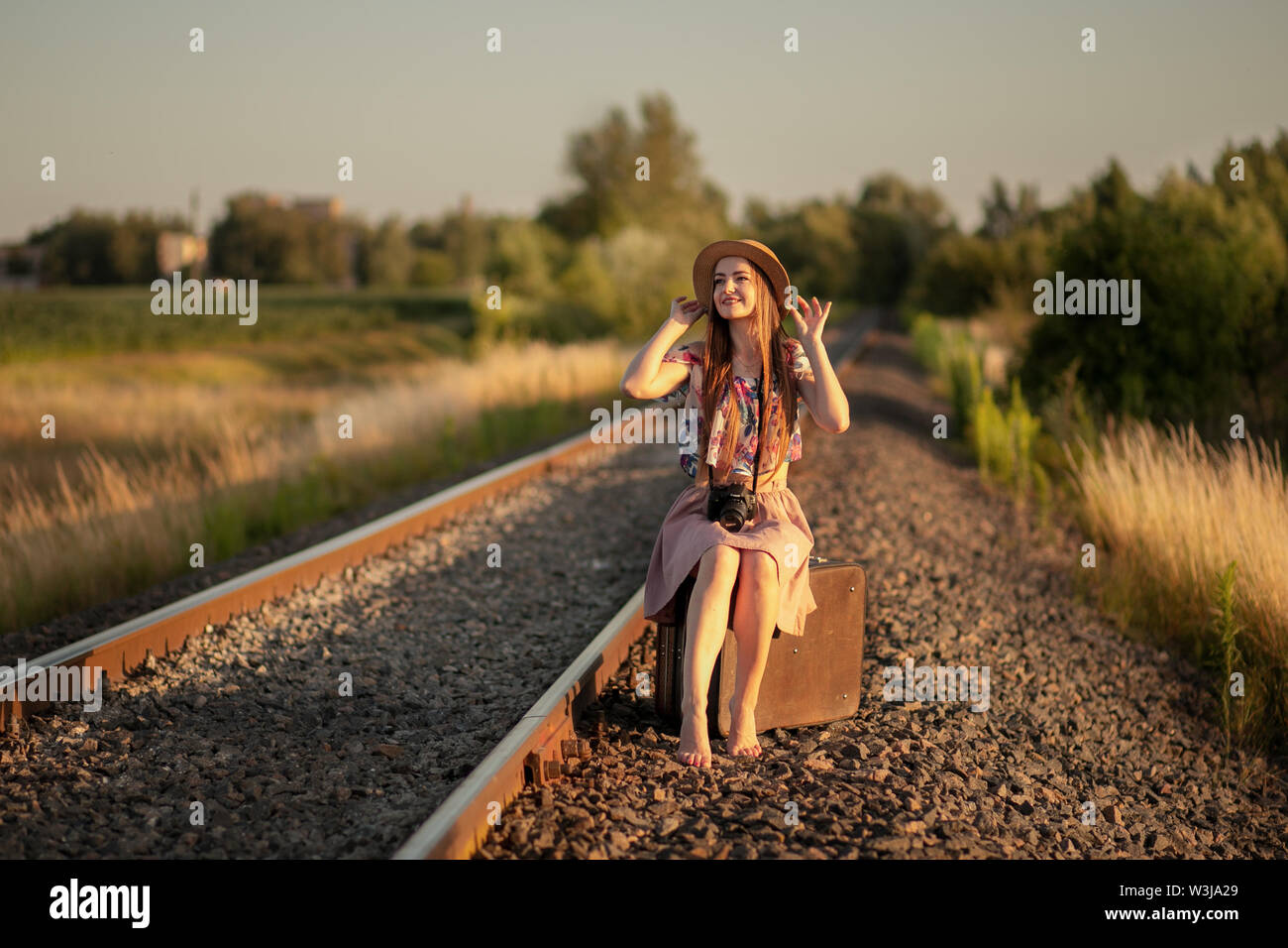 Slim ragazza in un cappello di paglia seduti sulle valigie sui binari della ferrovia con una telecamera, sorridente e sognare. Il concetto di viaggio, felicità, libertà, Foto Stock