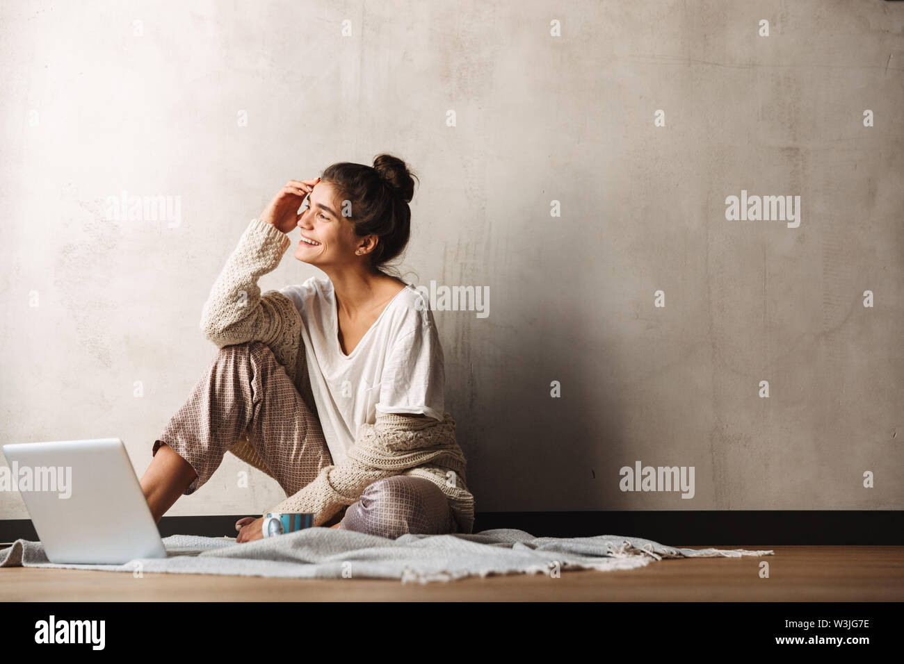 Foto di felice incantevole donna che indossa abiti per il tempo libero a bere caffè e utilizzando computer portatile mentre è seduto sul pavimento a casa Foto Stock