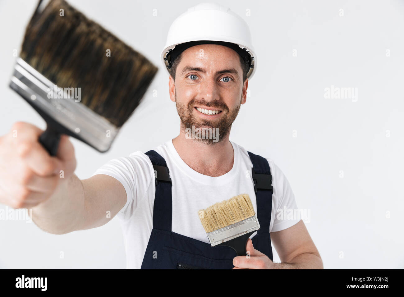Fiducioso barbuto builder uomo indossare tuta e casco in piedi isolato su sfondo bianco, shownig pennelli Foto Stock