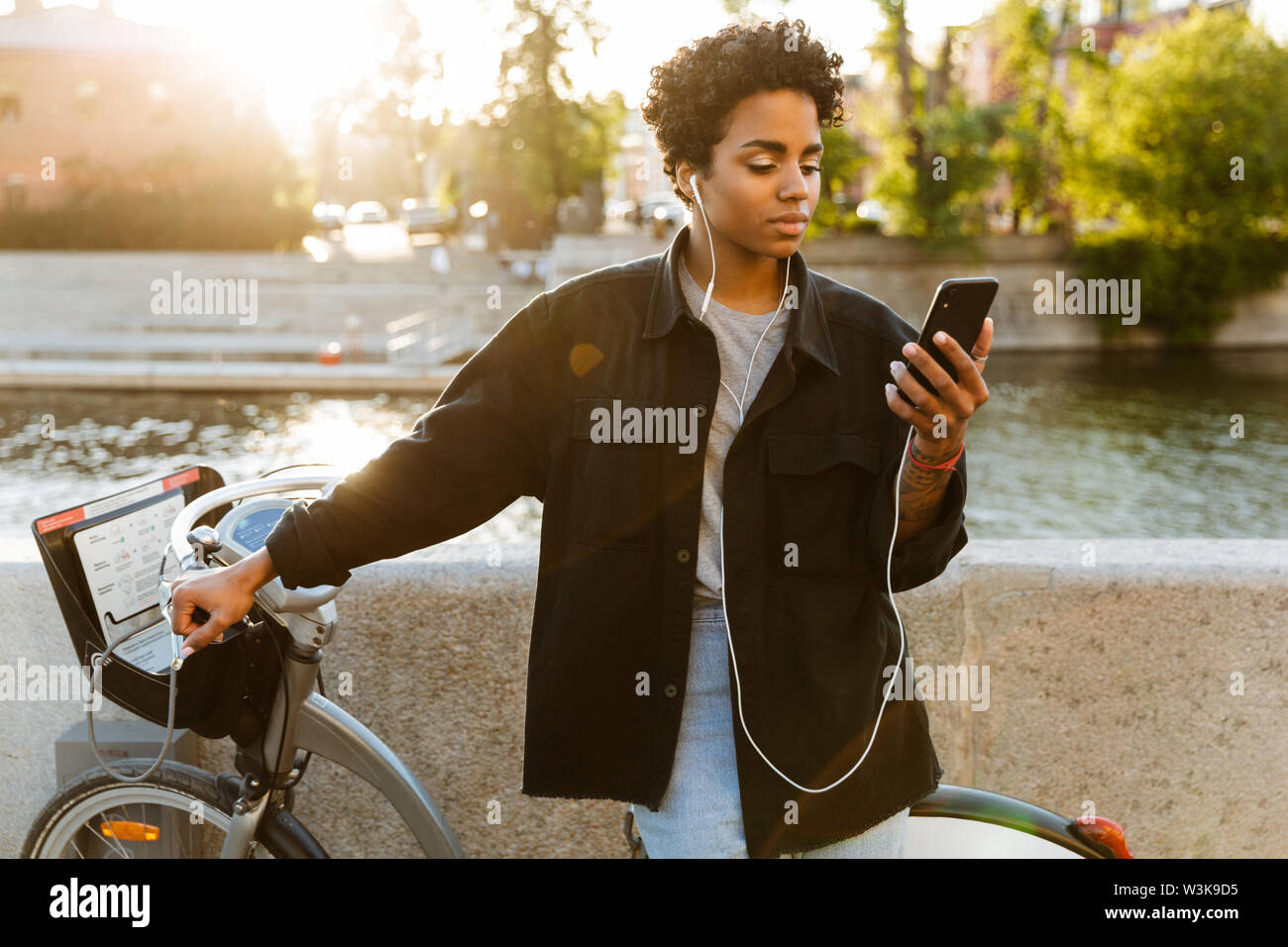 Foto di pensiero americano africano donna che indossa abiti casual in piedi con la bicicletta e utilizzando il cellulare mentre si cammina lungo il Riverfront Foto Stock