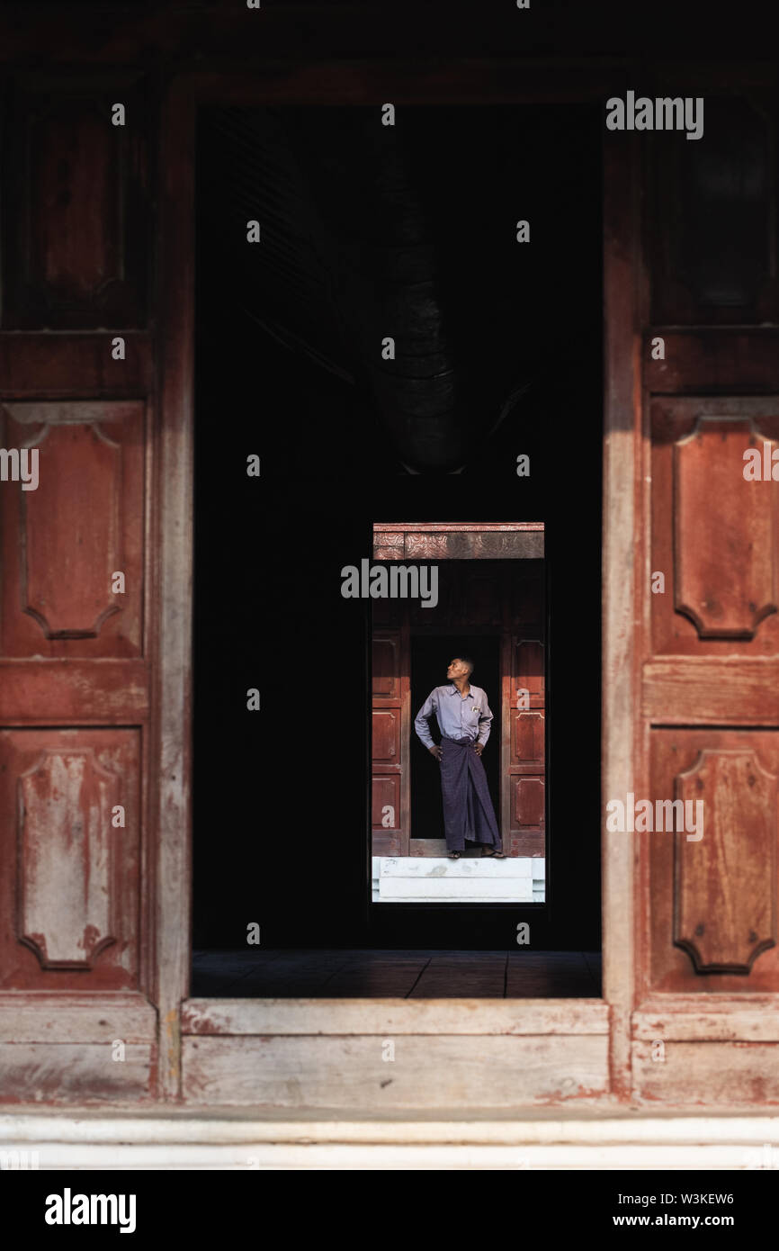 Mandalay, Myanmar - Aprile 2019: Burmese uomo in gonna longyi in piedi nel Palazzo Reale porta. Inquadratura con un arco in legno, con orientamento verticale Foto Stock