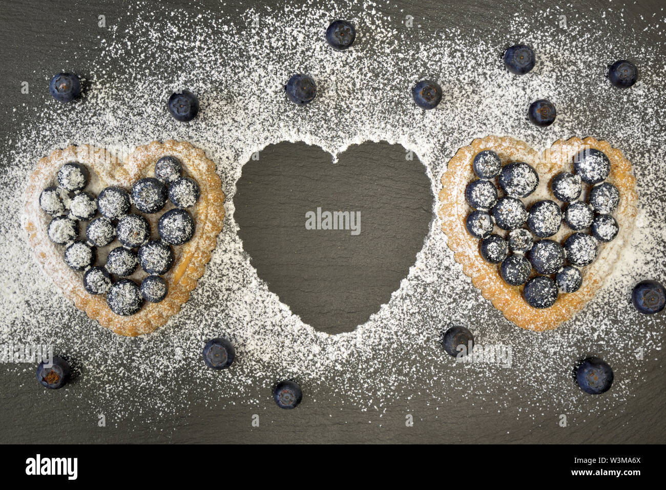 Due piccoli blu a forma di cuore torte cake giacciono su una piastra di ardesia. Essi sono spolverati con zucchero a velo e guarnita con mirtilli. Una torta è mancante Foto Stock