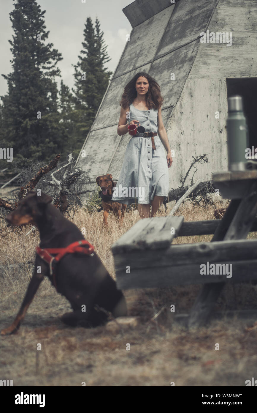 Un attraente brunette donna porta due tazze di caffè per una tavola mentre due cani sono intorno a lei. Yukon Territory, Canada Foto Stock