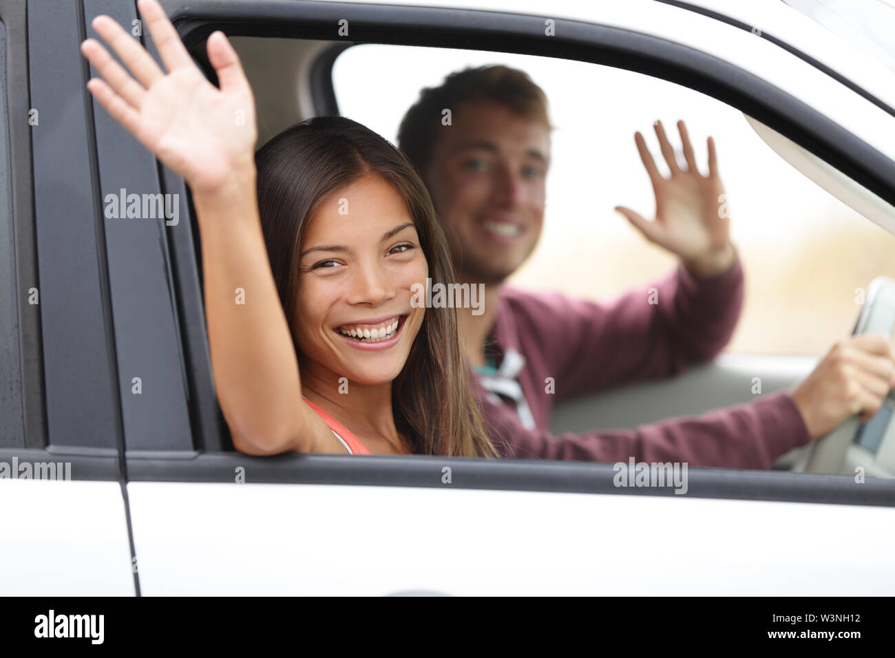 Driver guida in auto agitando felice a telecamera. Una giovane coppia in viaggio in auto nuova. Interracial coppia felice. Foto Stock