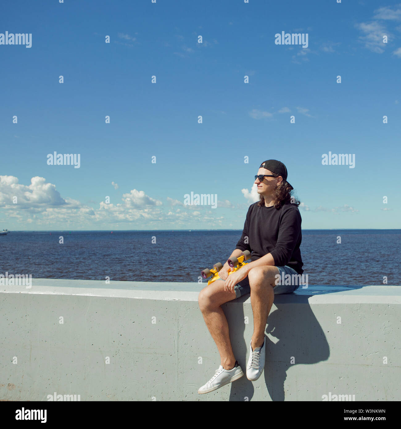 Un giovane uomo con capelli lunghi in un cappello da baseball e gli shorts in denim, felpa e occhiali neri è seduto con un pattino giallo sulla città's Quay. Copia dello spazio. Foto Stock