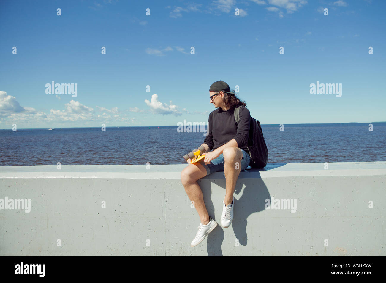 Un giovane uomo con capelli lunghi in un cappello da baseball e gli shorts in denim, felpa e occhiali neri è seduto con un pattino giallo sulla città's Quay. Copia dello spazio. Foto Stock