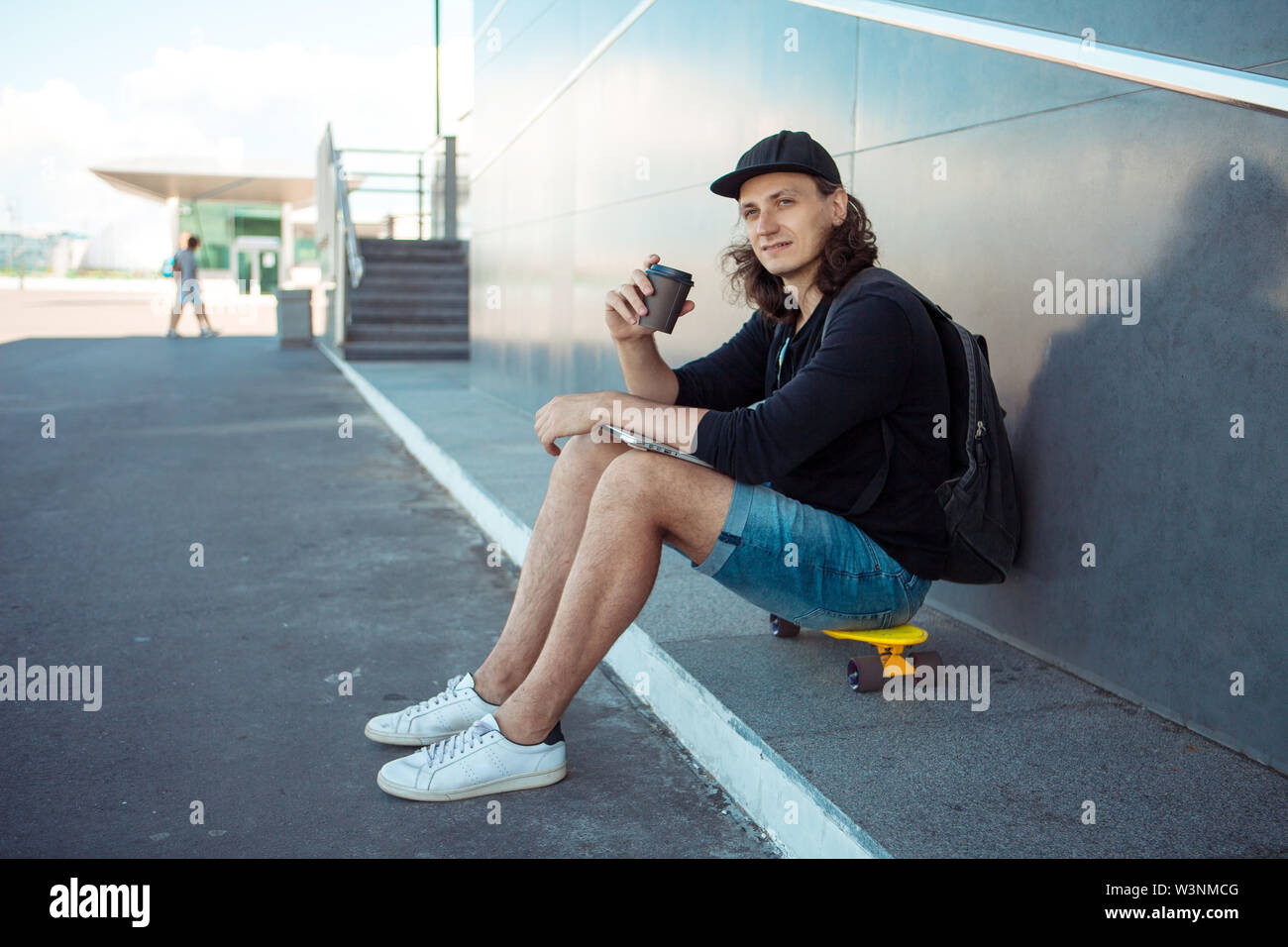 Un giovane uomo in un cappello da baseball, con uno zaino e gli shorts in denim, beve caffè seduto su uno skateboard giallo sull'asfalto. Foto Stock