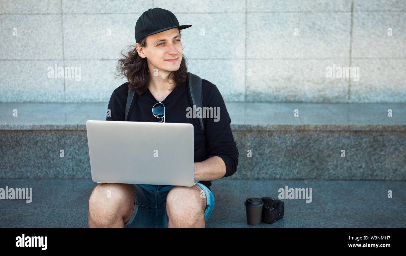 Il freelance che lavorano in aria aperta, concetto, giovane con capelli lunghi si siede sui gradini di granito in ombra, con un computer portatile. Estate giornata di vento. Copia sp Foto Stock