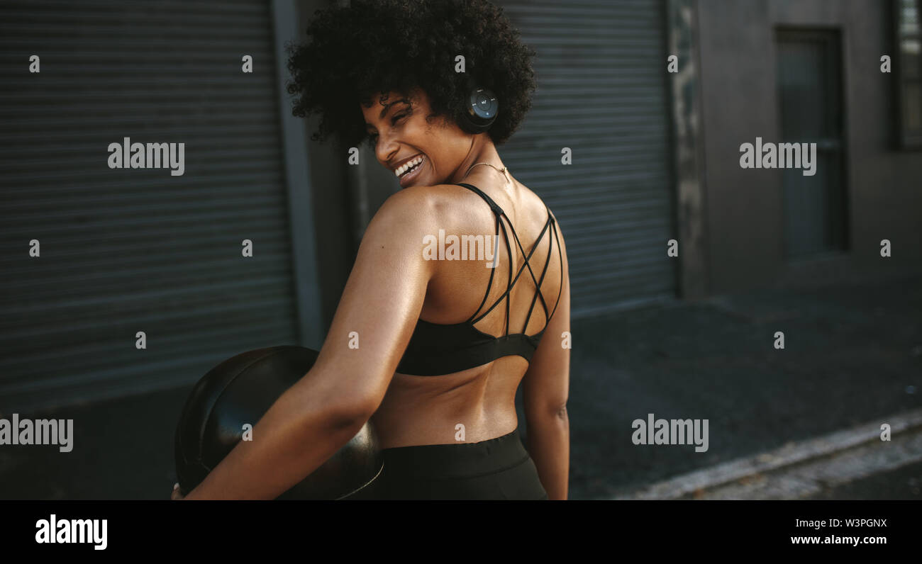 Vista posteriore della donna sorridente in usura fitness all'aperto permanente con la palla medica. Donna africana ascoltando musica su cuffie e riposo dopo wo Foto Stock