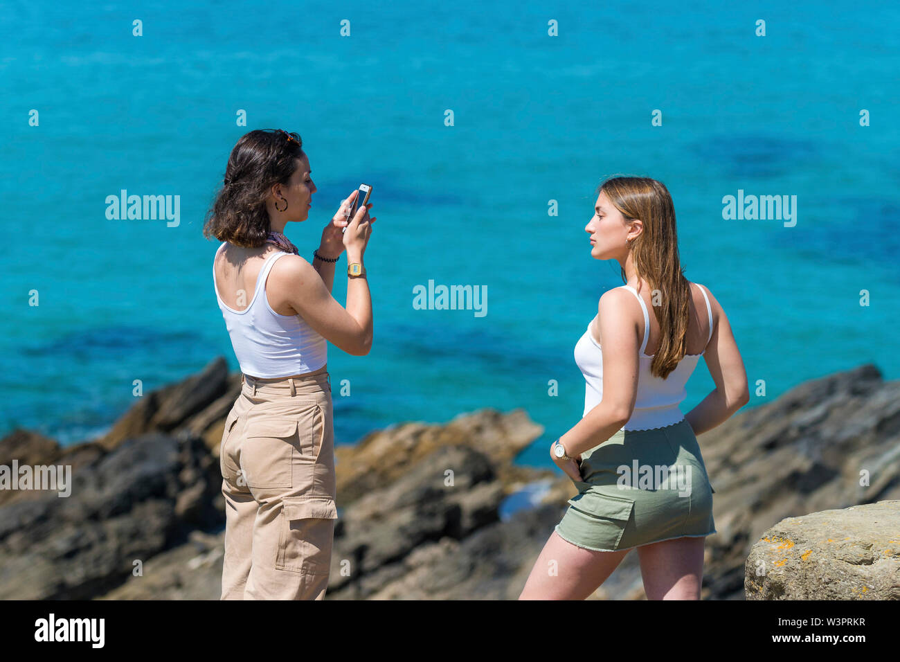 Una giovane donna di fotografare un altro giovane donna usando un telefono cellulare con un 'turqoise' mare colorato in background. Foto Stock