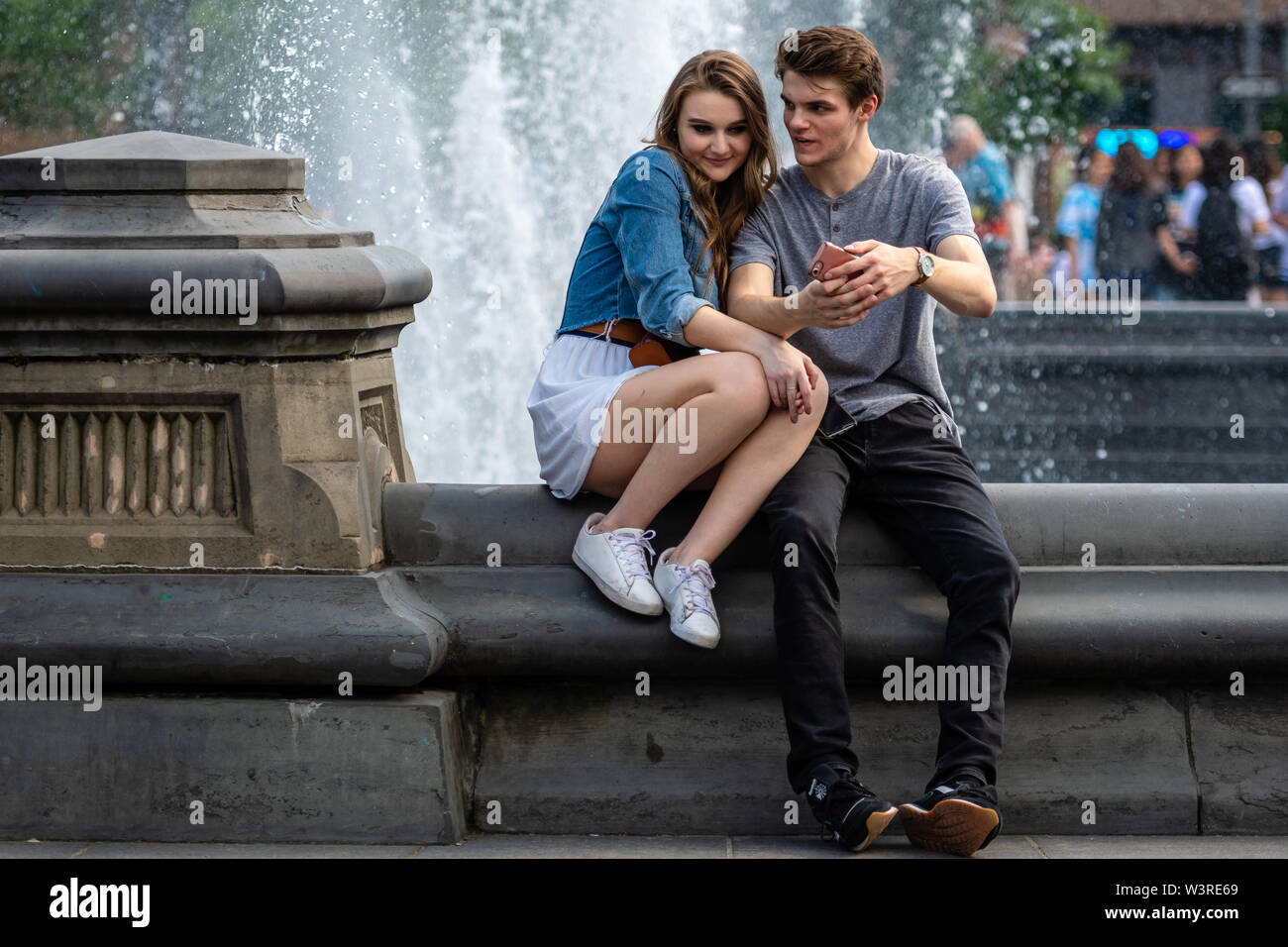 New York, Stati Uniti d'America - 21 Giugno 2019: una giovane coppia è seduto in un parco nei pressi di una fontana e parlando e guardando sul telefono. Foto Stock