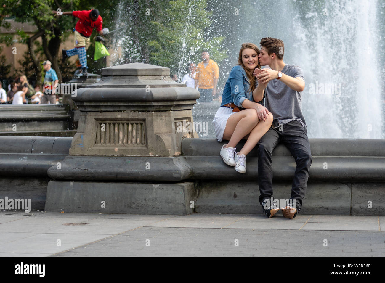 New York, Stati Uniti d'America - 21 Giugno 2019: una giovane coppia è seduto in un parco nei pressi di una fontana e parlando e guardando sul telefono. Foto Stock