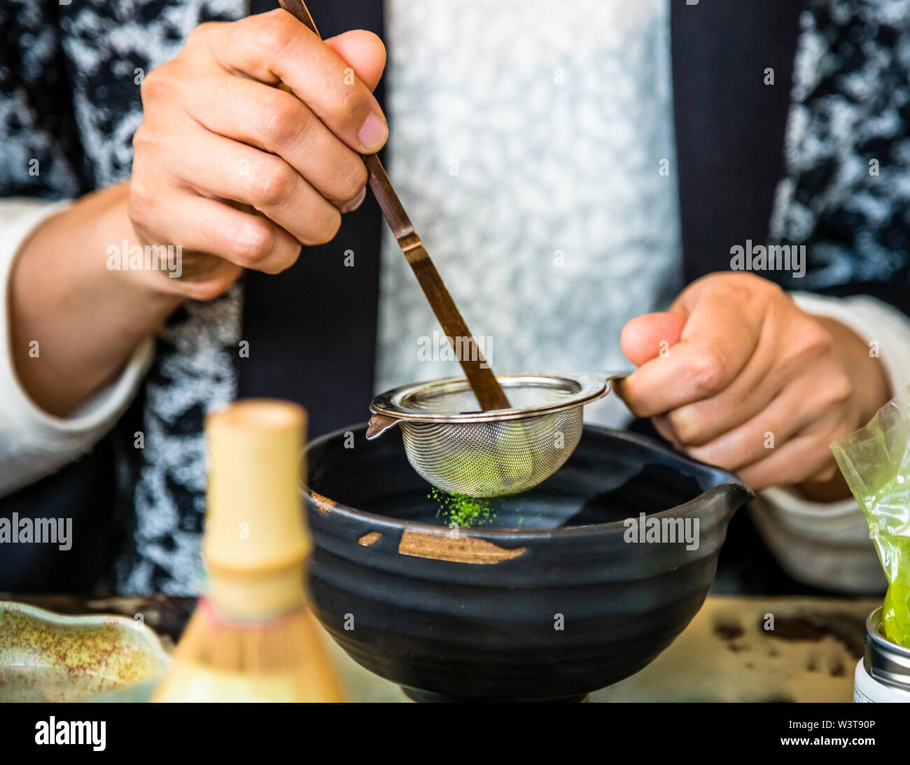 La maestra del tè Motoko Dobashi setaccia la polvere di matcha attraverso un setaccio fine usando un cucchiaio di bambù (chashaku) in una ciotola da tè scura durante una cerimonia del tè giapponese presso il negozio di tè ANMO a Düsseldorf, in Germania. Accanto si trova un contenitore d'argento con un secondo cucchiaio di bambù. Queste sono presumibilmente le mani della maestra del tè Motoko Dobashi. Foto Stock