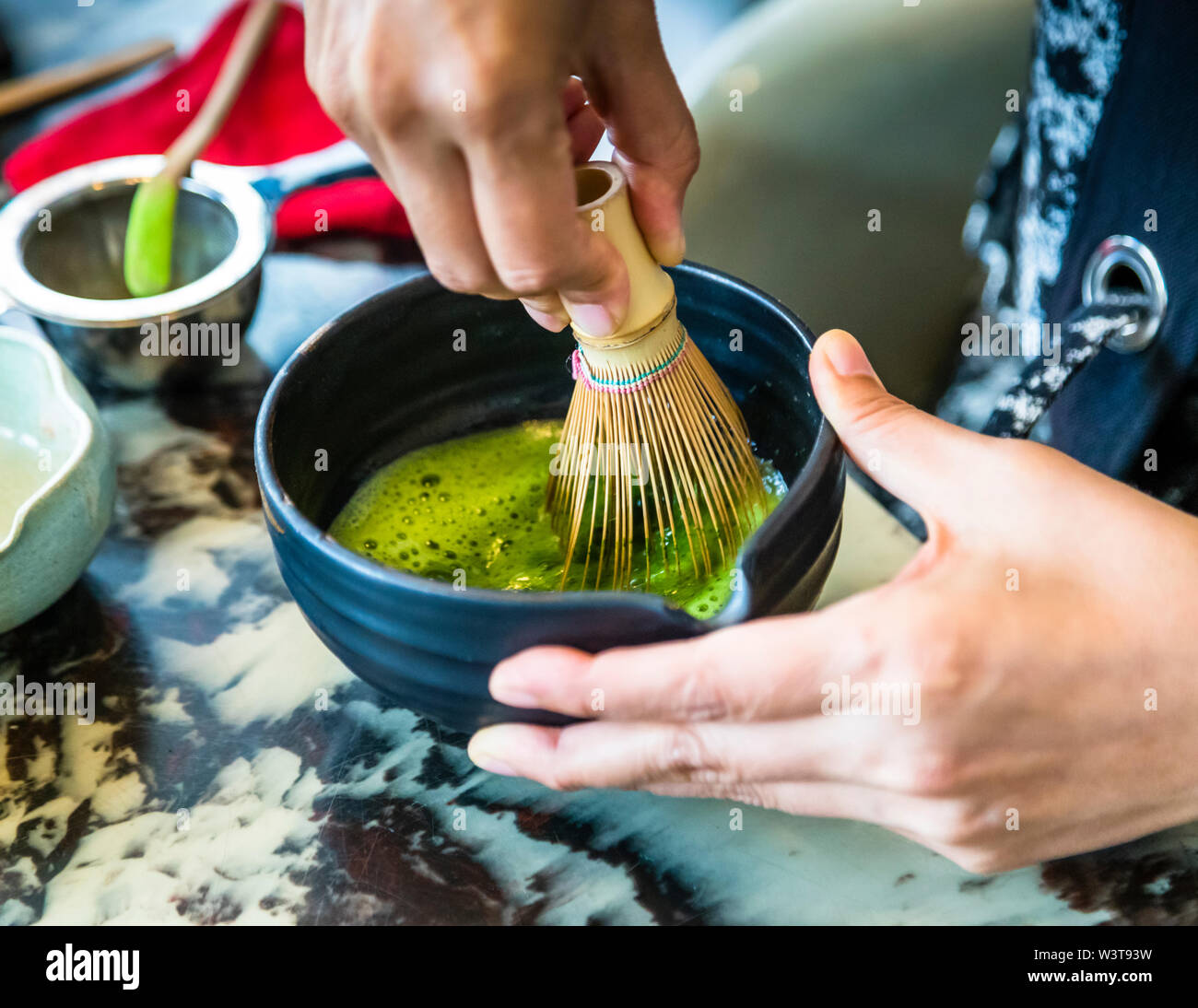 La maestra del tè Motoko Dobashi frusta il tè matcha in una schiuma verde usando un frullino di tè in bambù (chasen) in una ciotola da tè scura durante una cerimonia del tè giapponese al negozio di tè ANMO a Düsseldorf, Germania. Queste sono presumibilmente le mani della maestra del tè Motoko Dobashi. Foto Stock