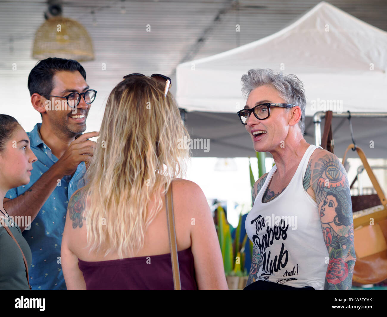 Un accoppiamento, pesantemente tatuato donna matura in bicchieri e serbatoio superiore conversa con persone a un 'Loca per locale' evento nel centro di Corpus Christi nel Texas, Stati Uniti d'America. Foto Stock