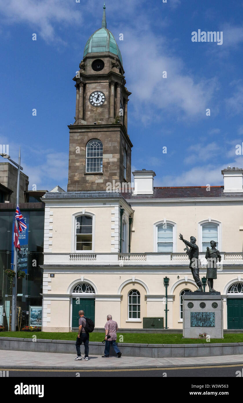 Due persone che camminano davanti al museo in piazza del mercato Lisburn, in Irlanda del Nord un pomeriggio estivo con un cielo blu dietro il museo guglia di clock. Foto Stock