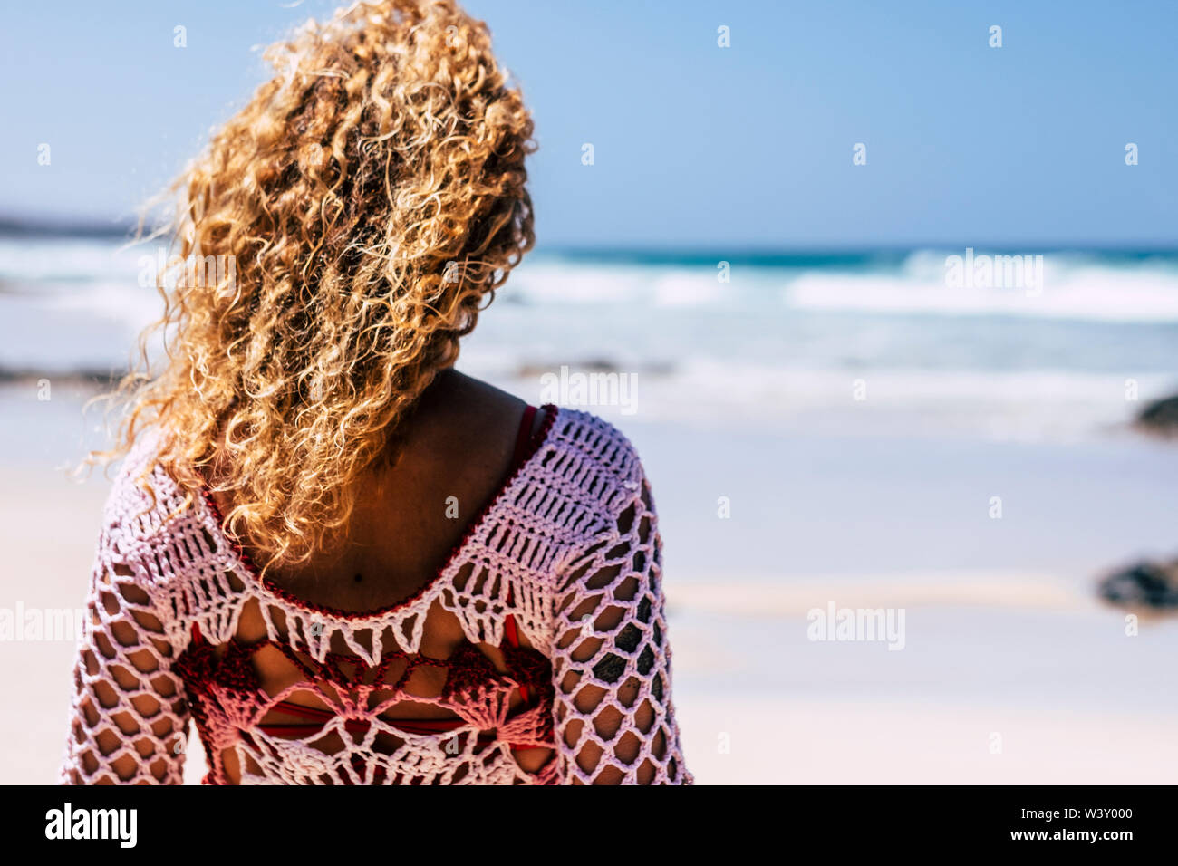 Blonde curly donna visto dal retro guardando il blu eocean onde in sfondo sfocato - Concetto di viaggi e vacanze estive per persone alla moda Foto Stock