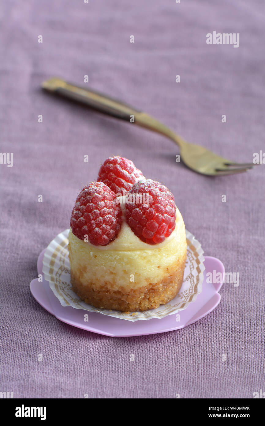 Deliziosi mini Cheesecake ai lamponi e rosa sul cupcake stand su viola pallido sfondo di lino. Il fuoco selettivo sulla parte anteriore di lamponi. Formato verticale. Foto Stock