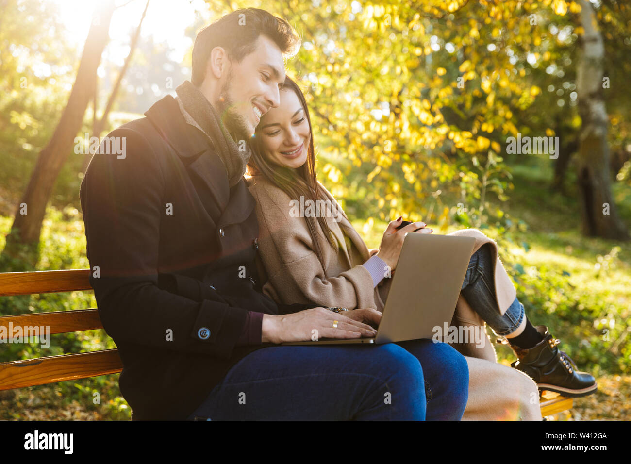 Allegro coppia giovane spendere tempo di divertimento presso il parco in autunno, seduta su una panchina, utilizzando laptop Foto Stock