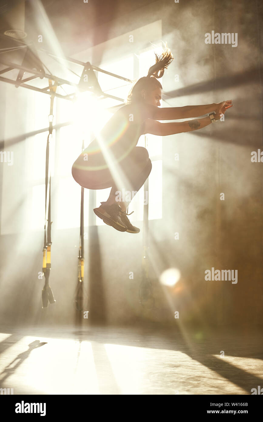 Superiore e superiore. Vista laterale della giovane donna atletica in sport abbigliamento jumping mentre ti alleni in palestra professionale. Sport concept. TRX Training. Allenamento Foto Stock