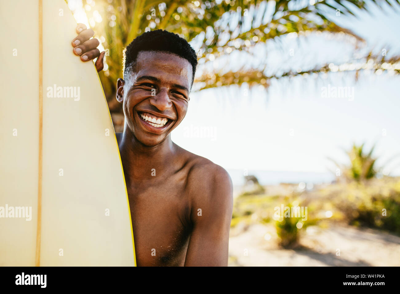 Sorridente giovane africano con la tavola da surf in piedi vicino a una spiaggia. Uomo sorridente con il surf board all'aperto su un giorno d'estate. Foto Stock