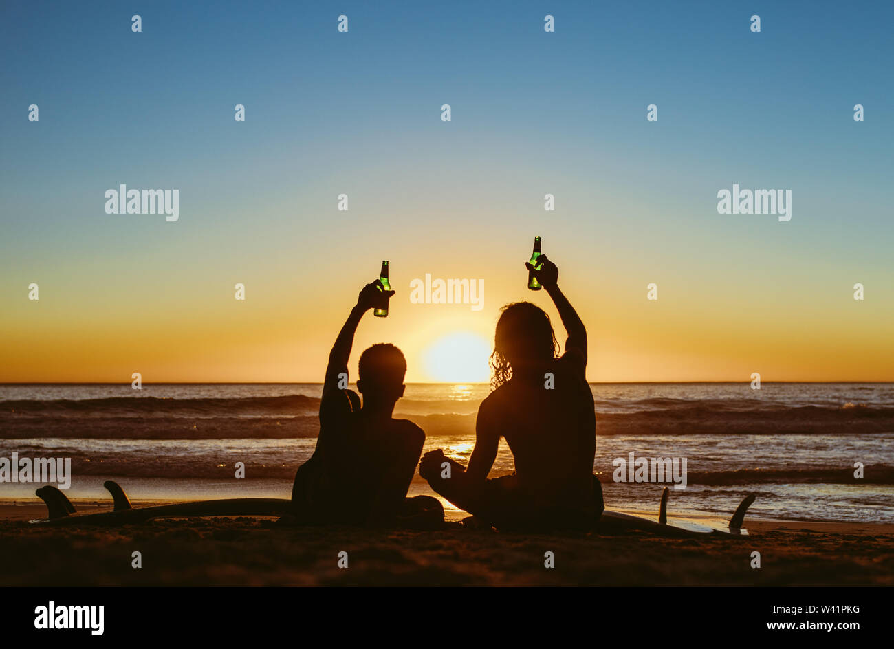 Vista posteriore di due giovani surfisti birre di tostatura sulla spiaggia e guardare il tramonto. Giovani uomini partying durante il tramonto sulla spiaggia. Foto Stock