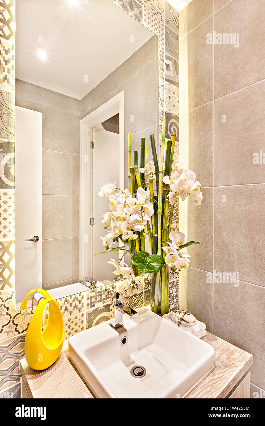 Fancy lavatoio con bambù e fiore bianco decorazione accanto a specchio con lavello e rubinetto, la parete dietro il vetro ha un modello di varie arti Foto Stock