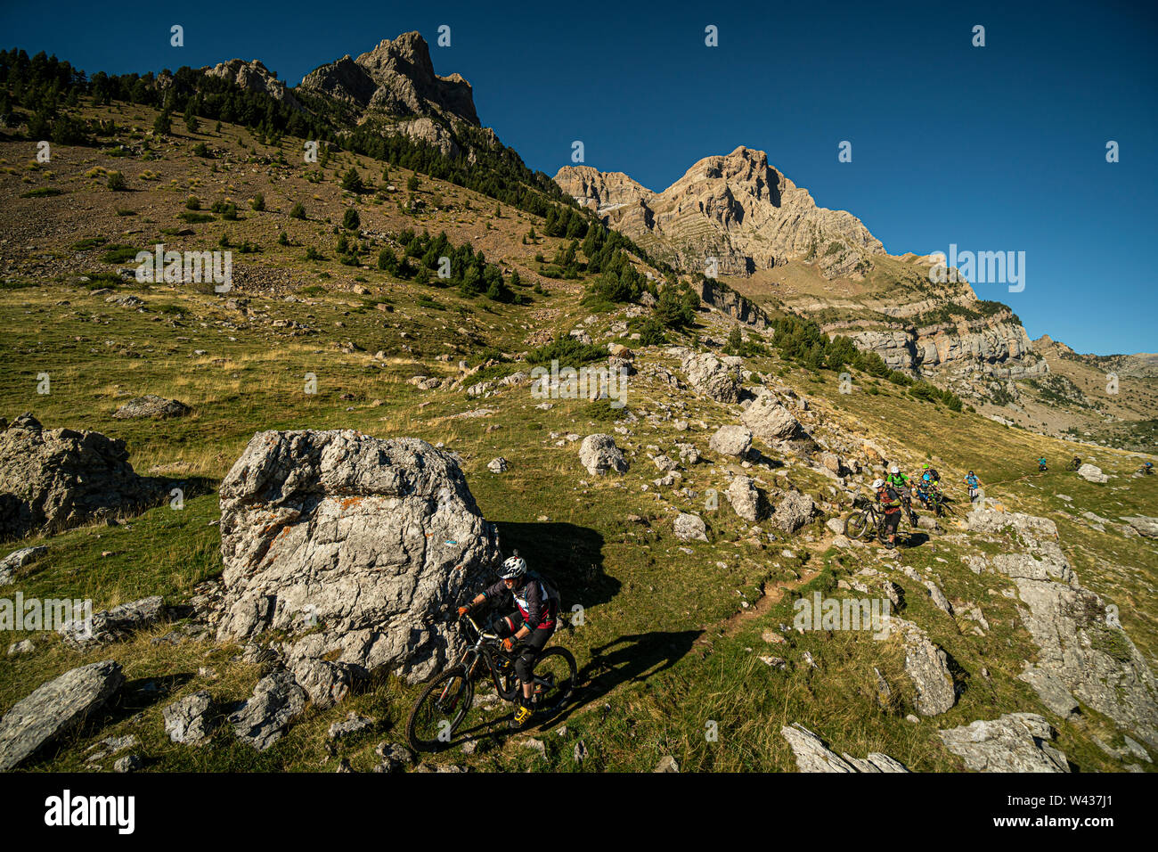 Un gruppo di appassionati di mountain bike a cavallo di un stretto sentiero roccioso con picchi drammatici e cielo blu dietro. Foto Stock
