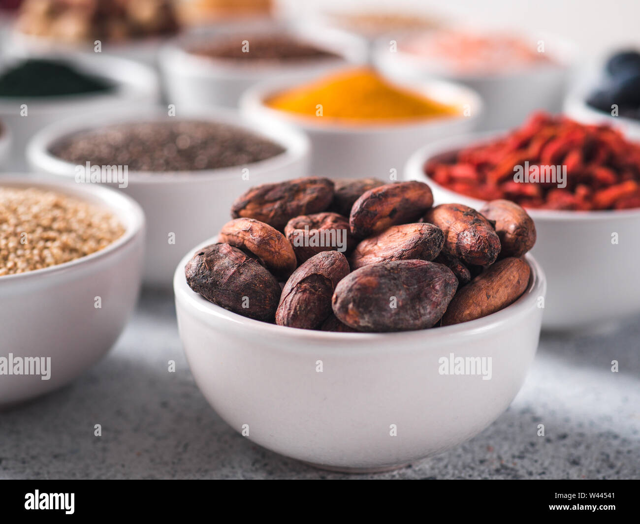 Materie fava di cacao in piccola ciotola bianco e altri superfoods sullo sfondo. Messa a fuoco selettiva. Superfoods diversi ingredienti. Concetto e illustrazione per la super e detox food Foto Stock